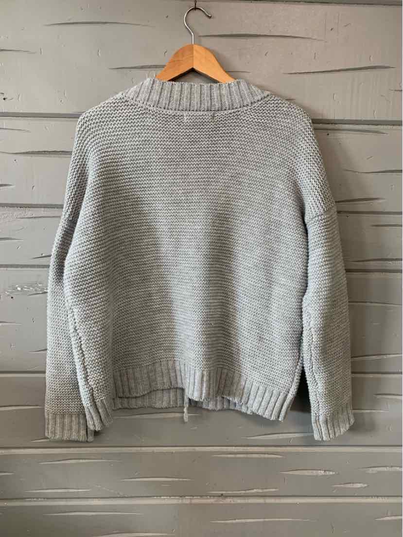Light gray knit sweater with ribbed collar, cuffs, and hem hanging on wooden hanger against gray wood panel wall.
