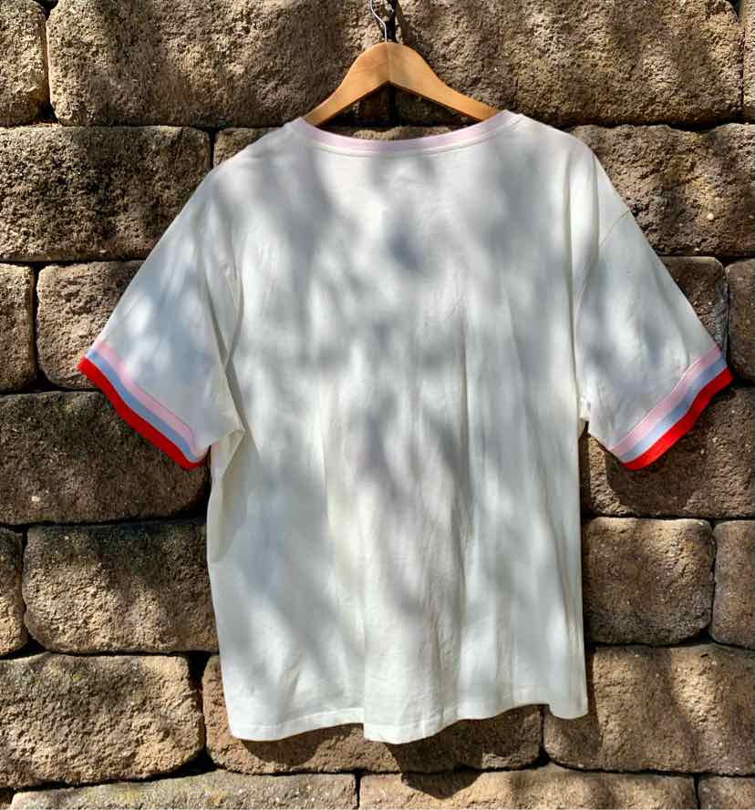 White short-sleeve t-shirt with red trim on cuffs, hanging against a stone wall background.