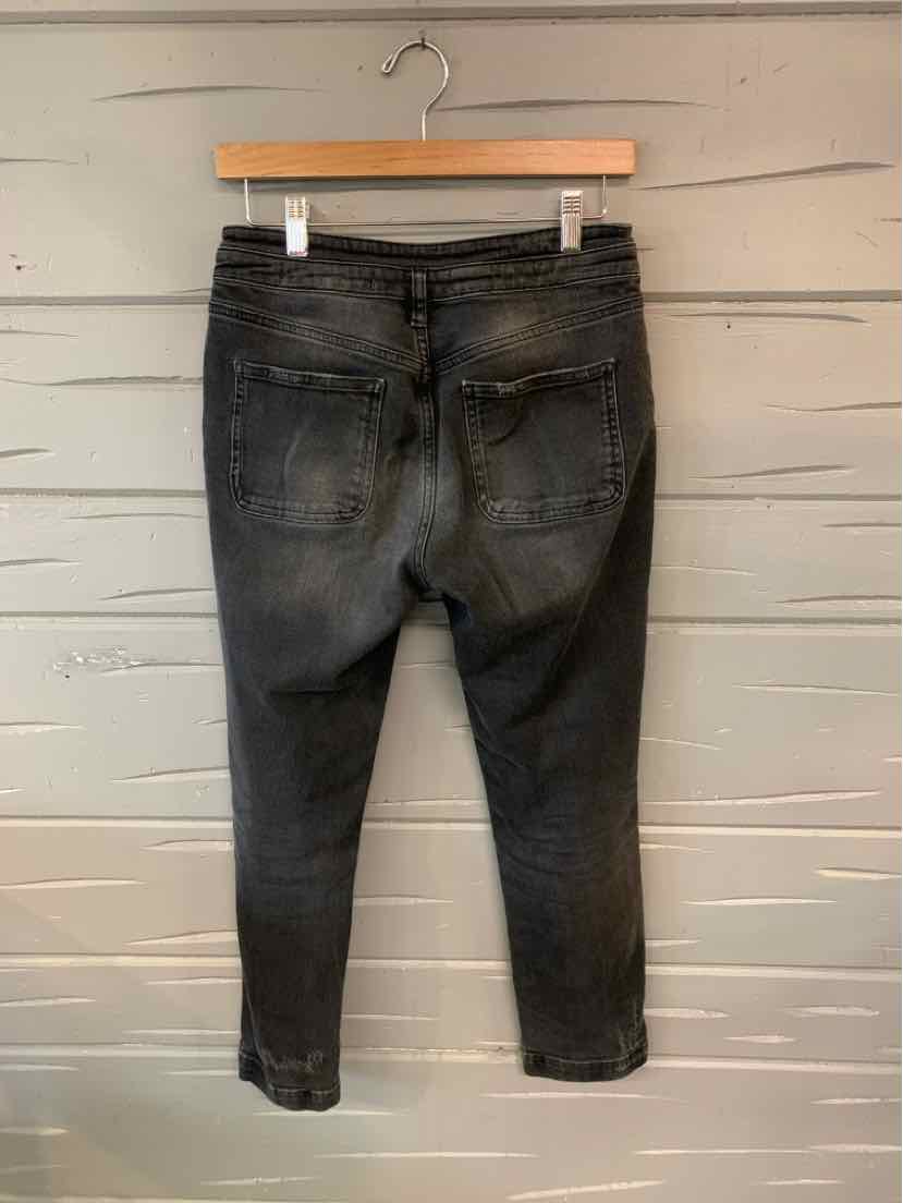 Black washed denim jeans with back pockets and tapered legs hanging on a wooden hanger against a light gray wooden wall.