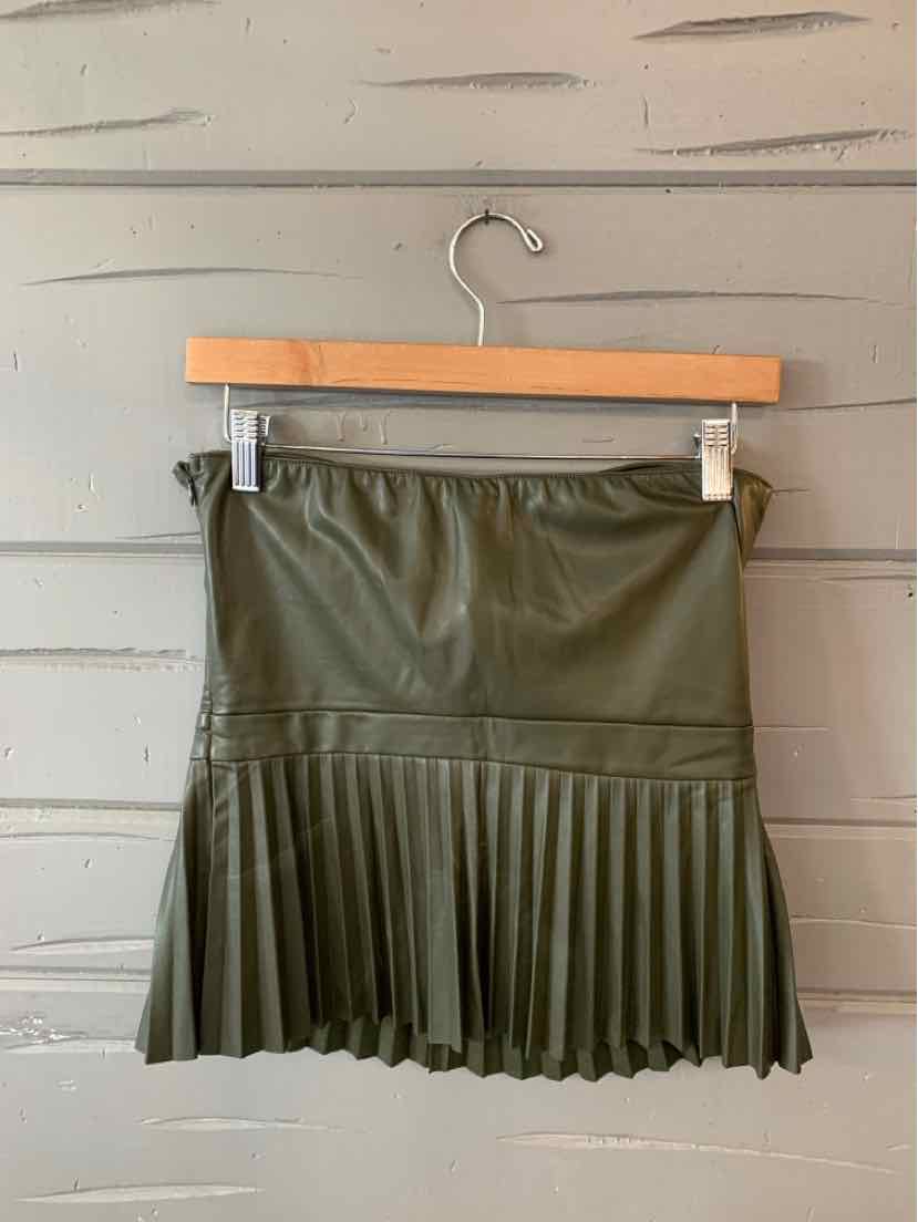 Green faux leather mini skirt with pleated hem hanging on wooden hanger against gray wooden wall.