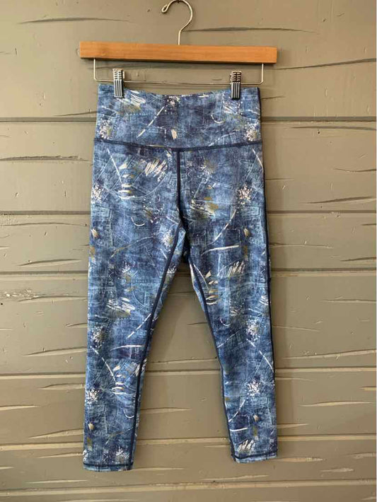 Blue tie-dye patterned leggings with high waistband hanging on wooden hanger against gray wooden wall.