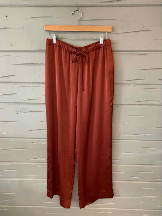 Brick red wide-leg pants with drawstring waist hanging on wooden hanger against rustic white wooden wall.