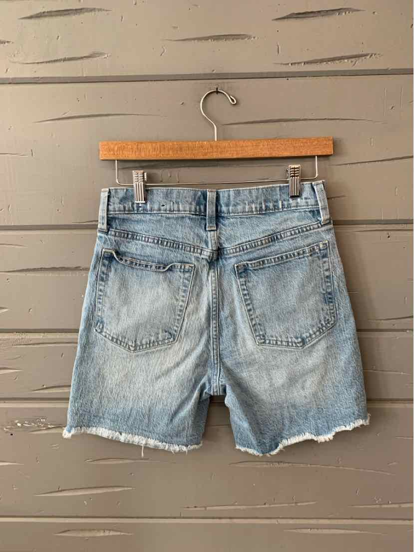 Light blue denim shorts with frayed hems and two back pockets hanging on wooden hanger against gray wooden wall.