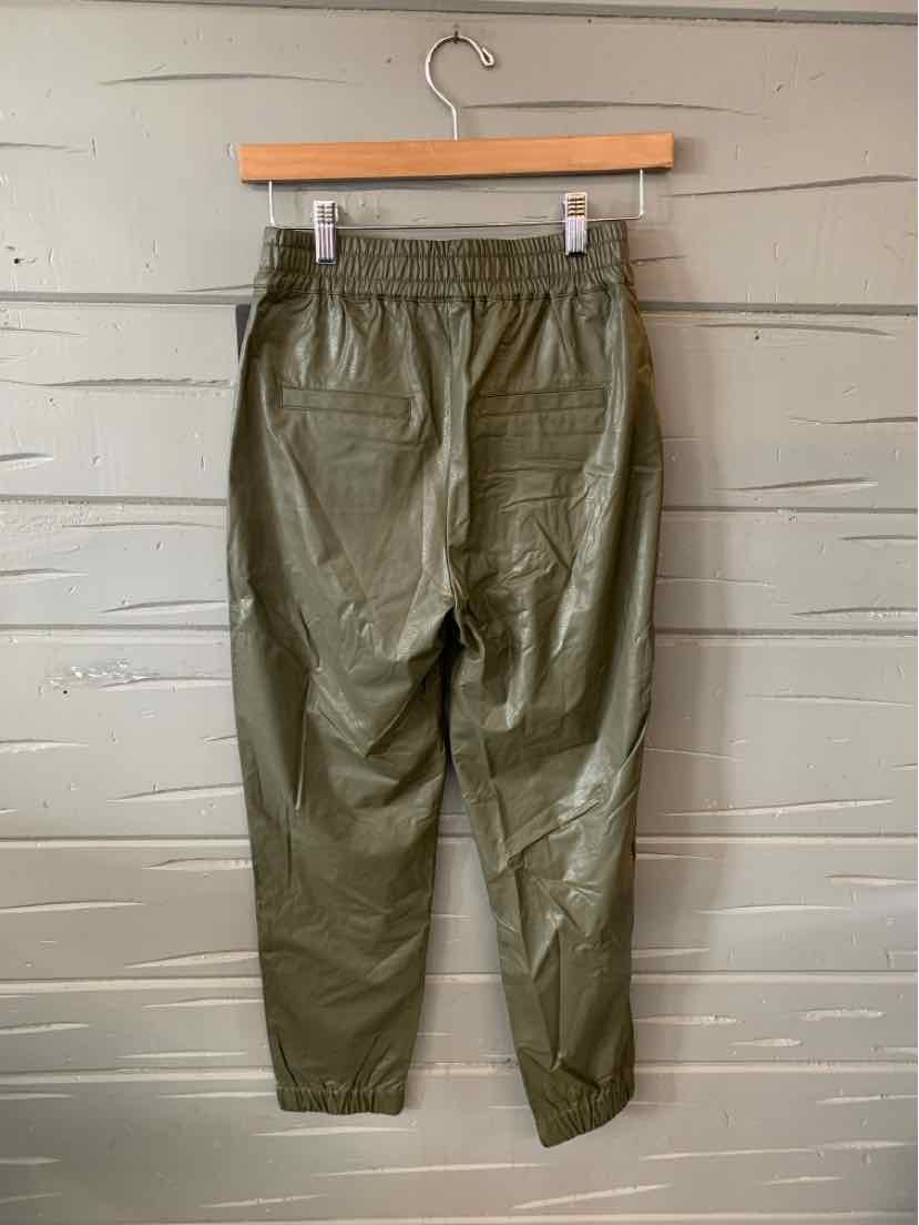 Olive green tapered pants with elastic waistband and ankle cuffs hanging on wooden hanger against gray wooden wall.