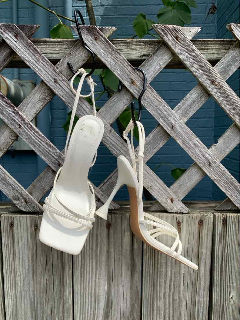 White strappy high-heeled sandals with square toes and thin ankle straps hanging on wooden lattice fence.