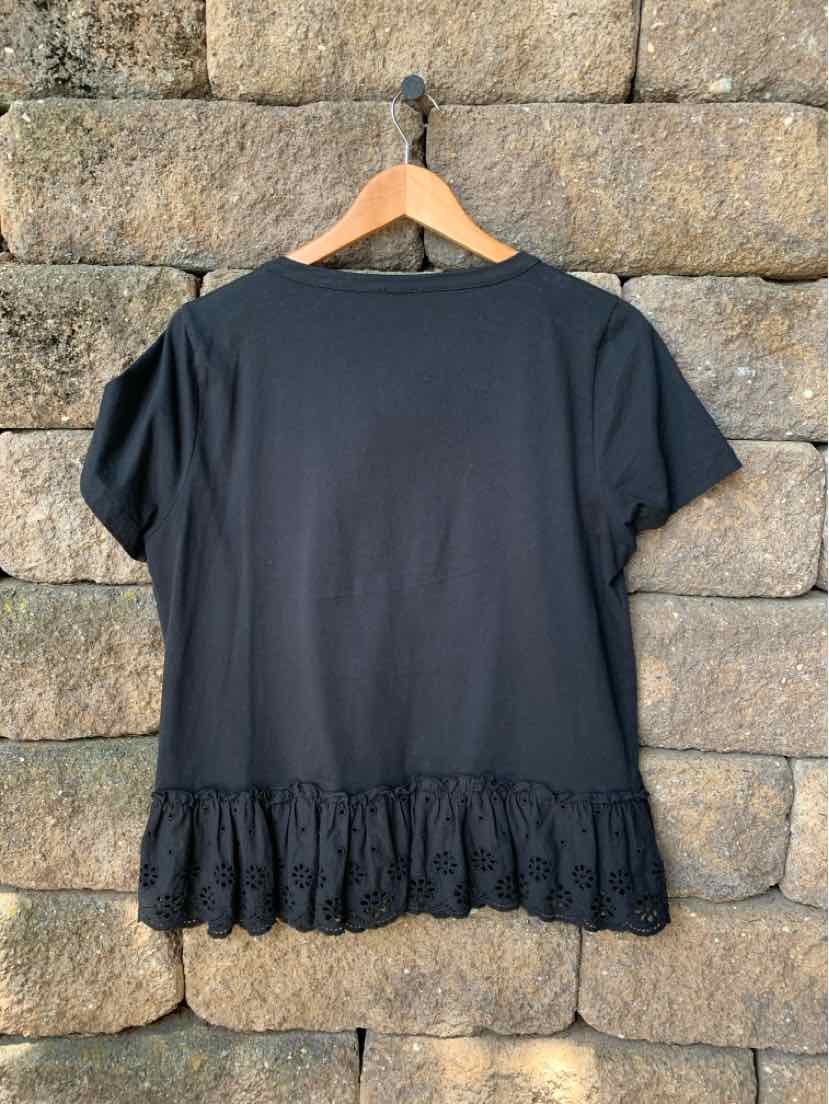 Black short-sleeve top with ruffled hem detail hanging on wooden hanger against stone wall.