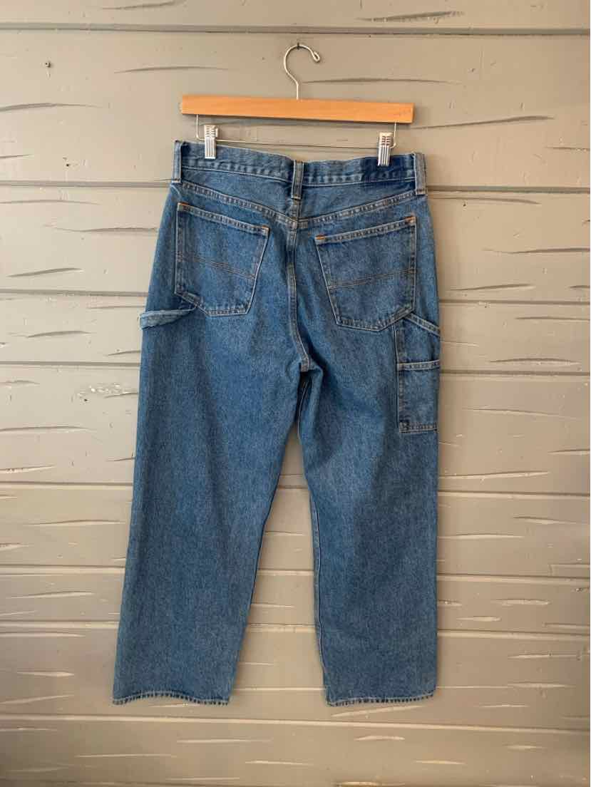 Blue denim workwear jeans with back pockets and side tool loop hanging on wooden hanger against beige wood wall.