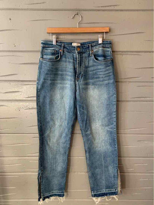 Blue washed denim jeans with front pockets and ankle zippers hanging on wooden hanger against gray wooden wall.