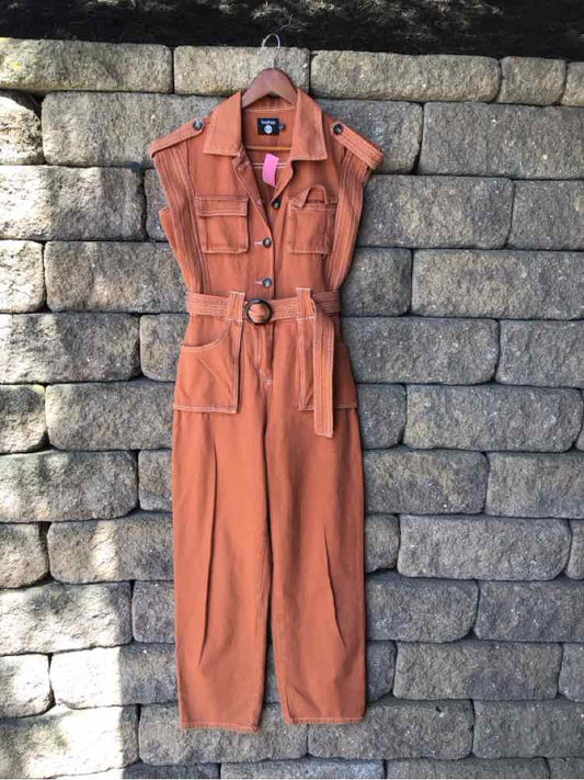 Rust orange sleeveless utility jumpsuit with belted waist and front pockets hanging against stone wall.