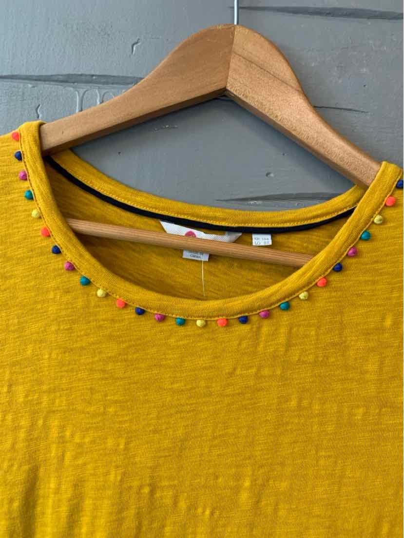 Mustard yellow textured fabric top with multicolored pom-pom trim around round neckline on wooden hanger.