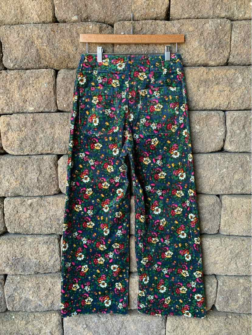 Dark floral patterned wide-leg pants with a high waist and button closure, hanging against a stone wall background.