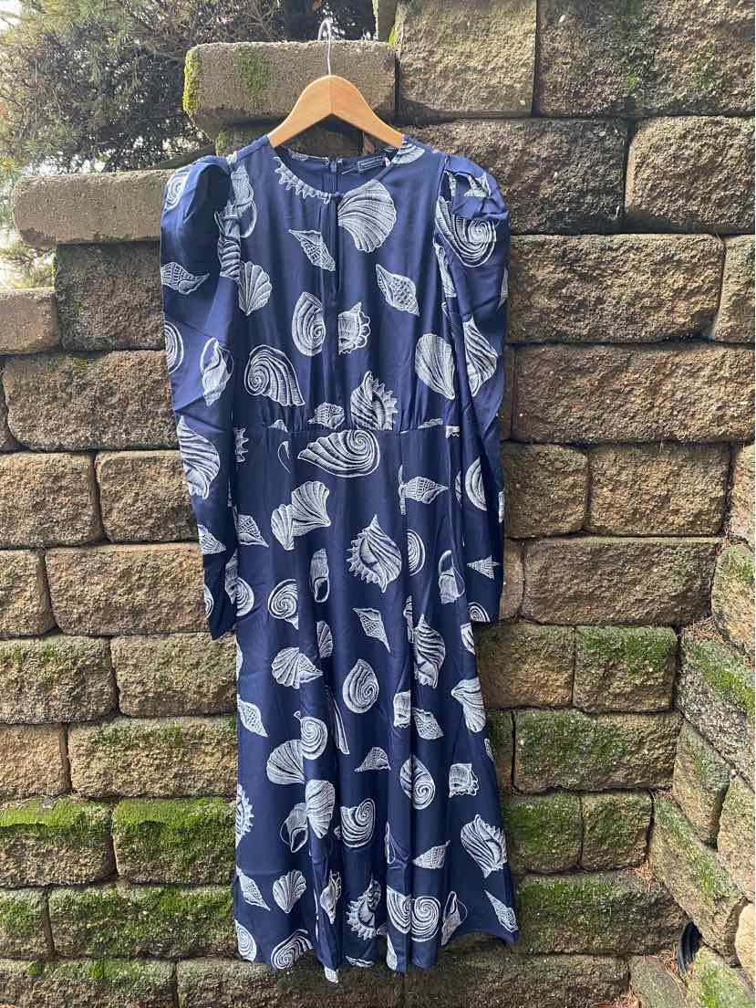 Navy blue maxi dress with white seashell print, long sleeves, and gathered waist hanging outdoors on wooden hanger.