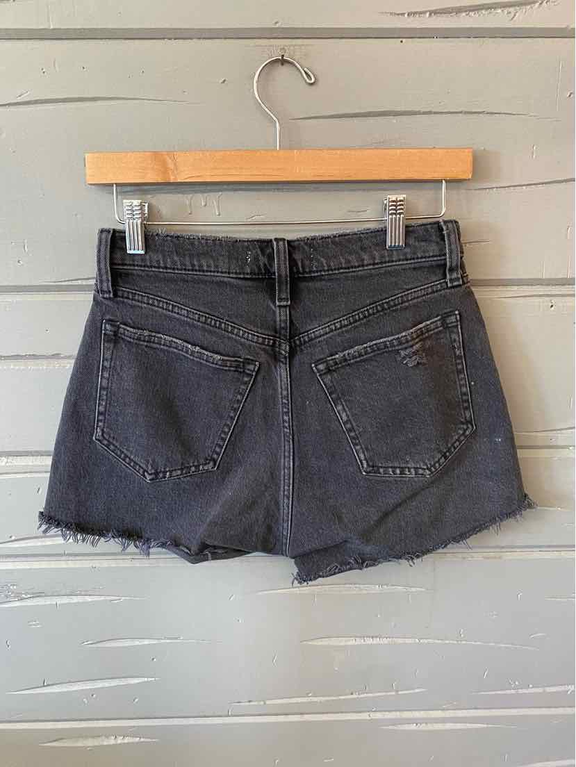 Black denim shorts with raw hem and back pockets on wooden hanger against gray wall.