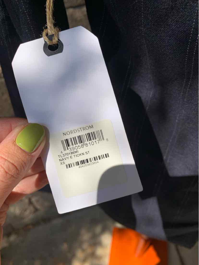 White rectangular clothing tag with black barcode and text, attached to black fabric, held by hand with green nail polish.