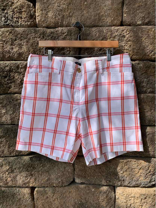 White shorts with red plaid pattern, knee-length, button and zipper closure, hanging on wooden hanger against stone wall.