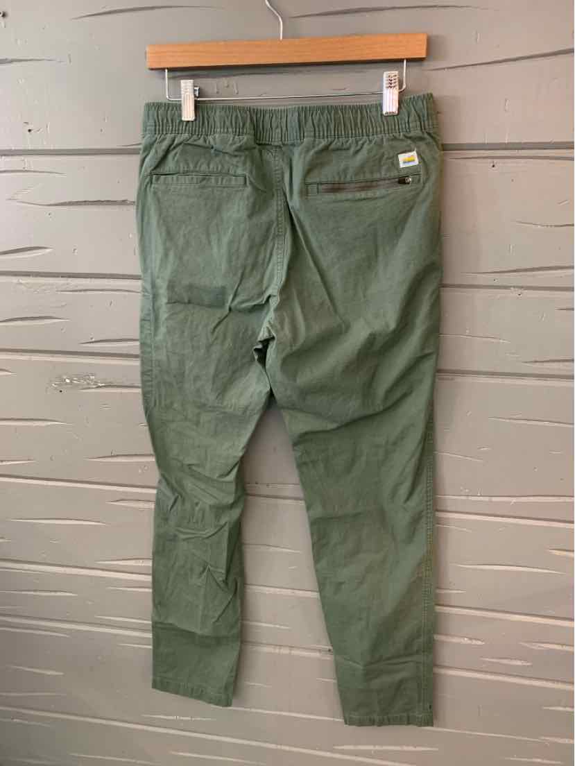 Green tapered pants with elastic waistband and two back pockets on wooden hanger against gray wall.
