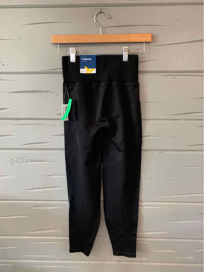 Black high-waisted leggings with a fitted ankle and tag details hanging on wooden hanger against gray wood wall.