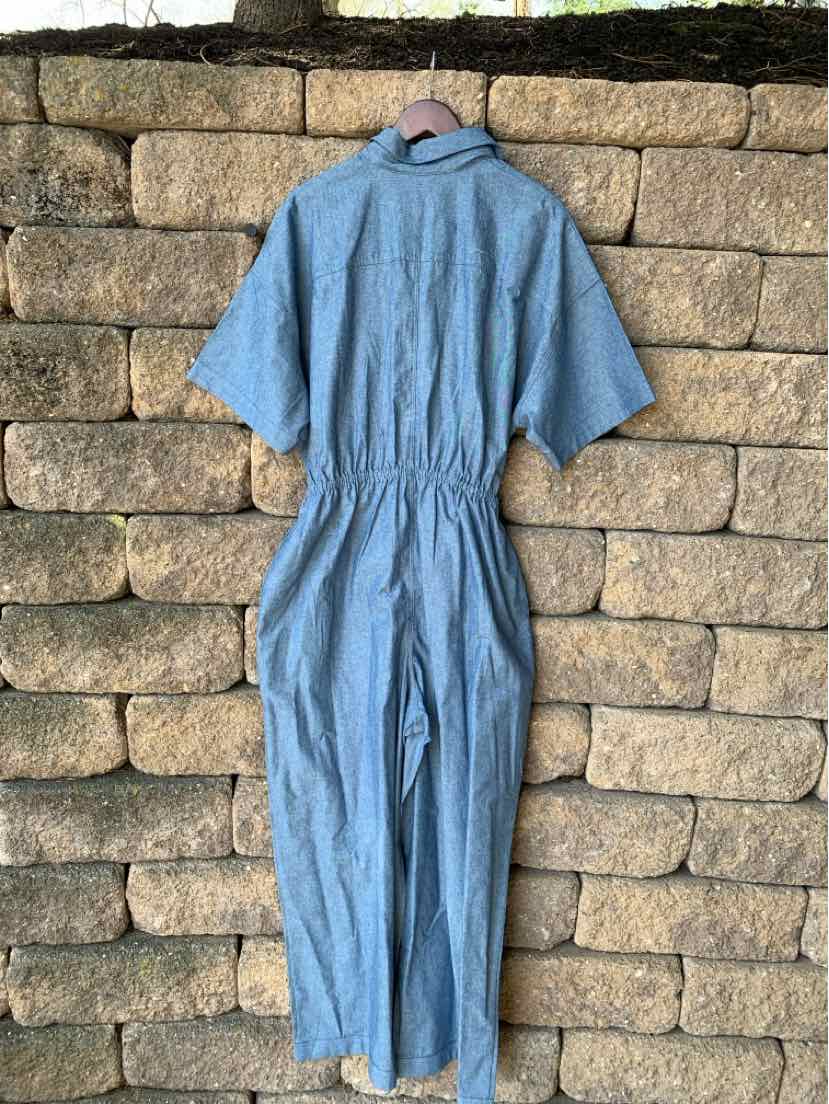Light blue short-sleeve jumpsuit with cinched waist and back zipper hanging against stone wall.