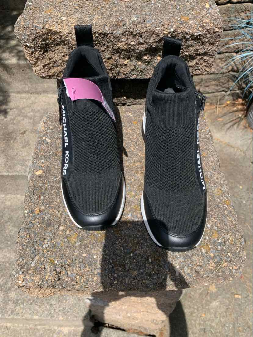 Black slip-on sneakers with mesh upper, rubber sole, and pull tabs; one shoe has a purple accent and Michael Kors logo on.