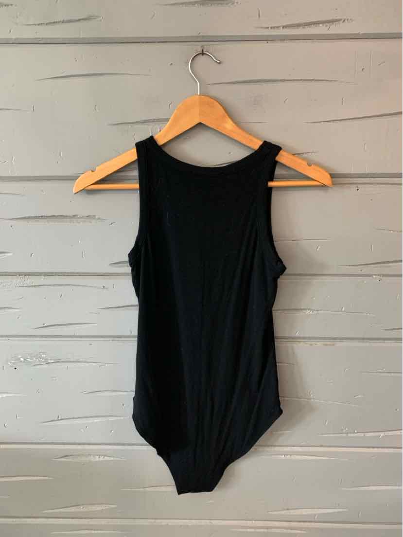 Black sleeveless bodysuit with scoop neckline hanging on wooden hanger against gray wall.