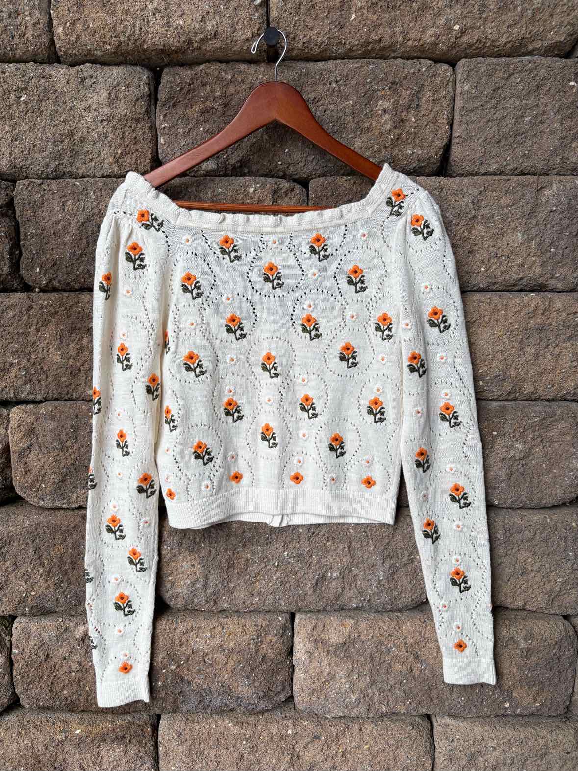 White cropped long-sleeve sweater with small orange and green floral embroidery and a wide neckline.