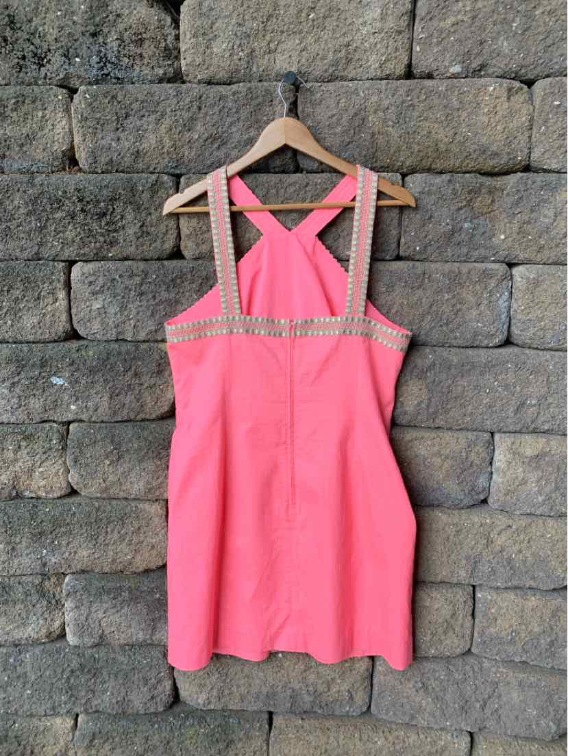 Coral pink sleeveless dress with crisscross straps and sequin trim on a wood hanger against stone wall.