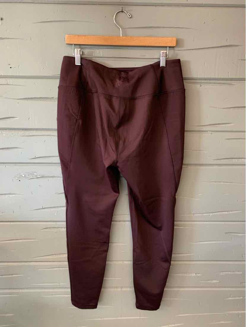 Dark burgundy tapered pants with elastic waistband and side pockets hanging on wooden hanger against gray wooden wall.