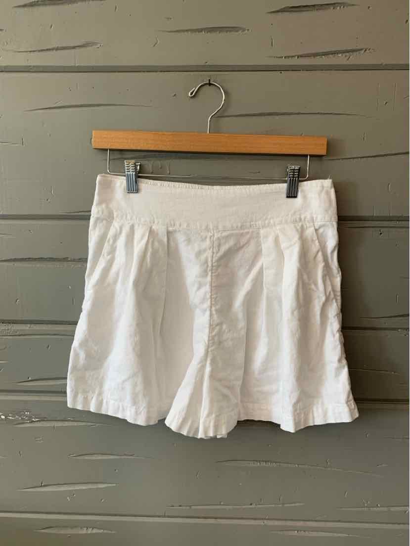 White high-waist shorts with front pleats hanging on wooden hanger against gray wooden wall.