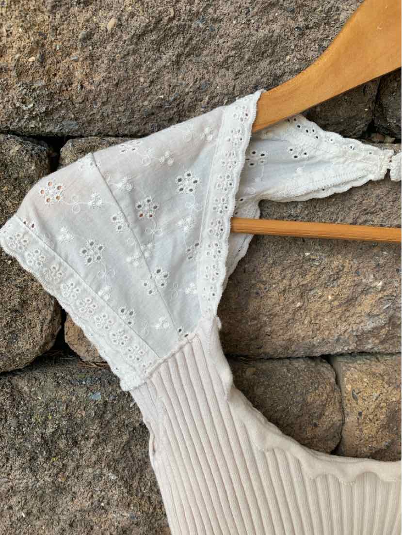 White eyelet lace cropped top with wide straps hanging on wooden hanger against stone wall.