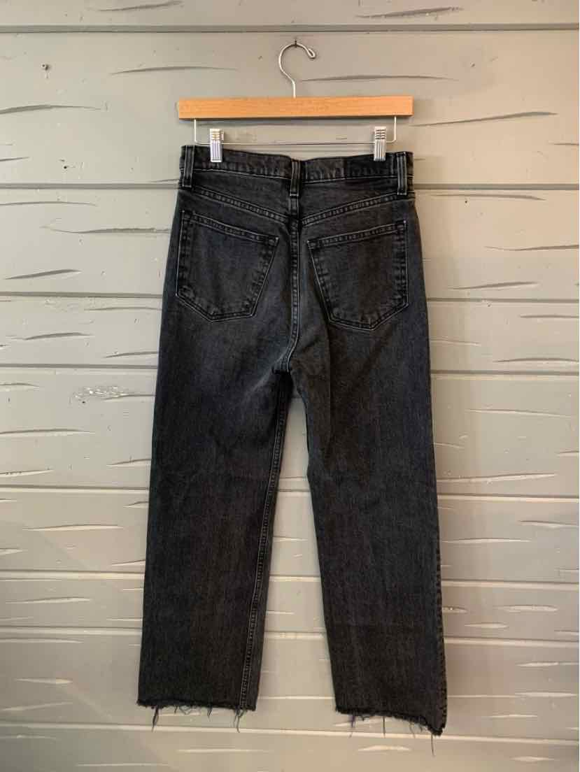 Black denim jeans with high waist, straight leg, frayed hems, and back pockets on wooden hanger against gray wall.