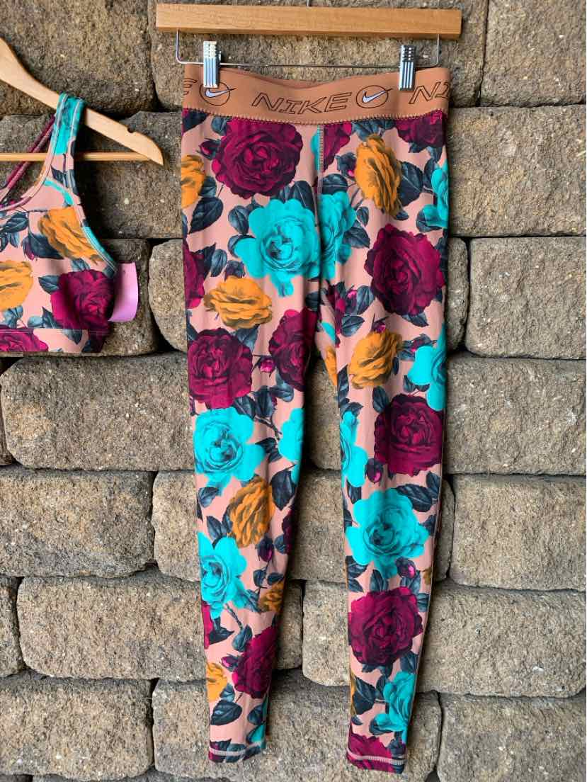 Floral patterned leggings in pink, teal, and mustard with brown waistband, hanging on wood hanger against stone wall.