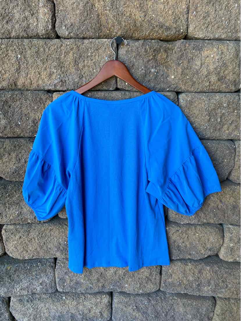 Bright blue blouse with balloon sleeves and a loose, boxy fit hanging on wooden hanger.