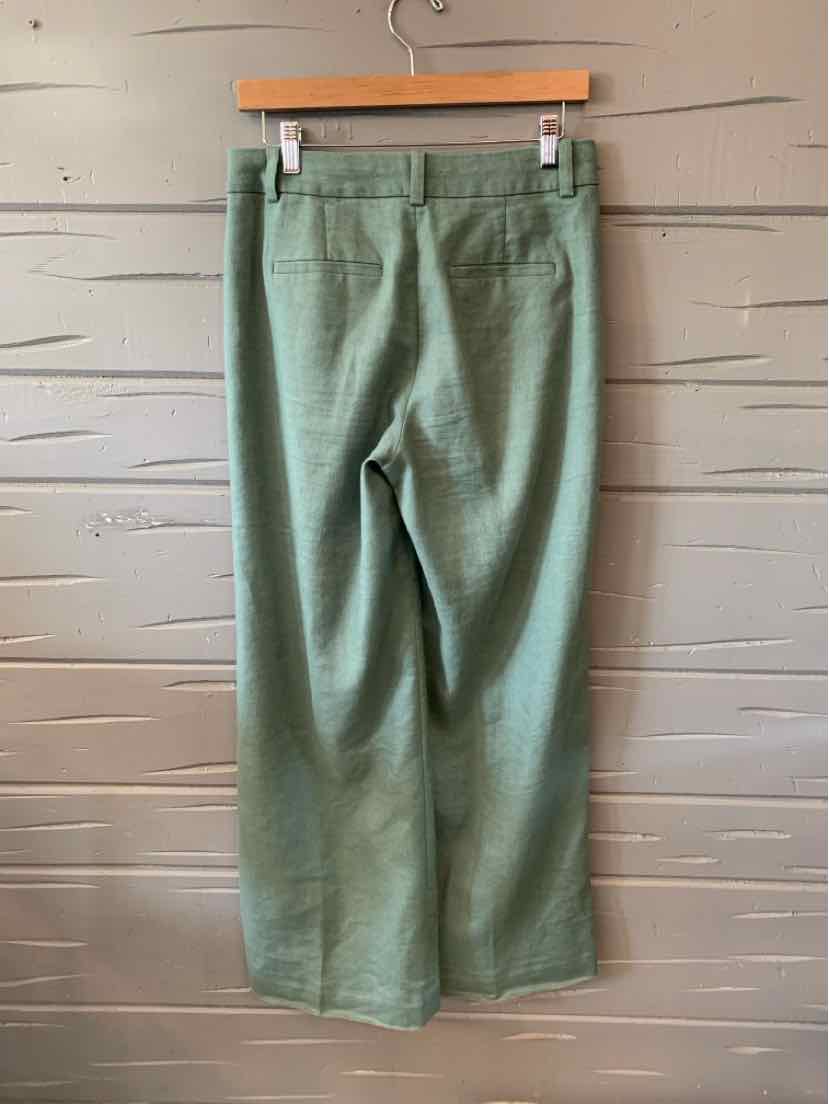 Light green straight-leg pants with back welt pockets hanging on wooden hanger against textured wall.