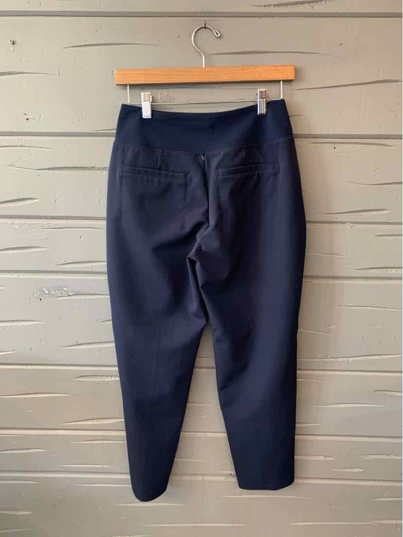 Navy blue tapered pants with elastic waistband and rear patch pockets hanging on wooden hanger against gray wooden wall.