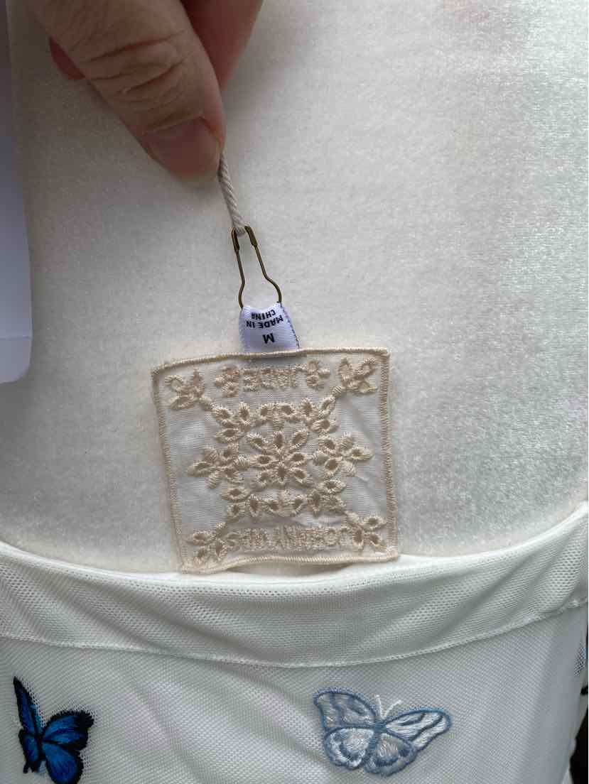 White fabric with beige floral embroidery patch and blue butterfly designs on pocket edge.