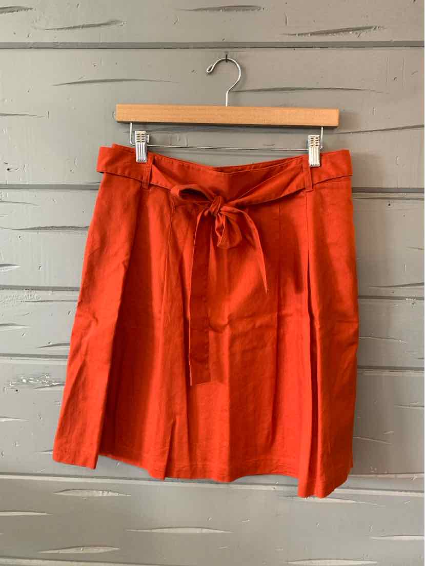 Rust-orange knee-length skirt with a waist tie belt and soft pleats hanging on wooden hanger.