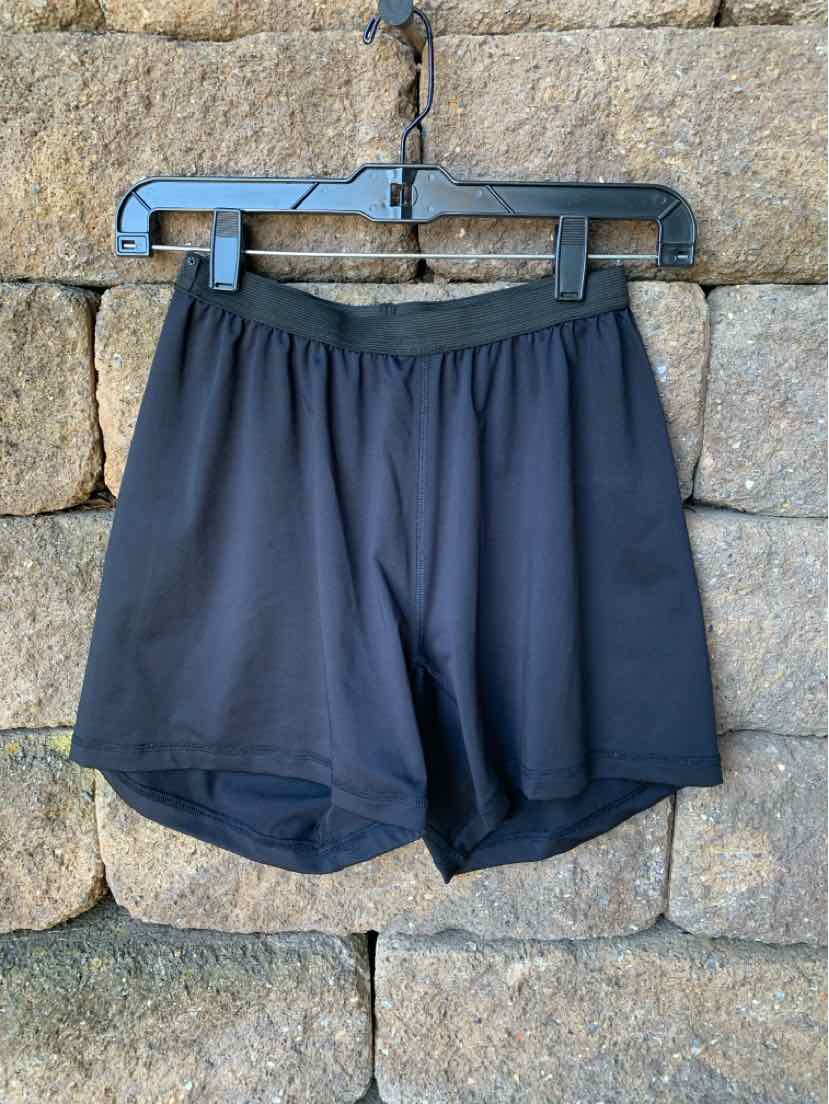 Black athletic shorts with elastic waistband, lightweight fabric, medium size, hanging on black hanger against stone wall.