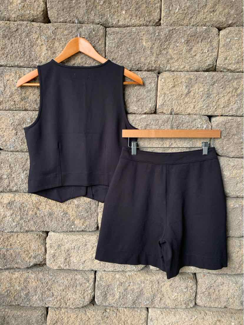 Black sleeveless top with round neckline and matching black shorts with a relaxed fit on wooden hangers against a stone wall.