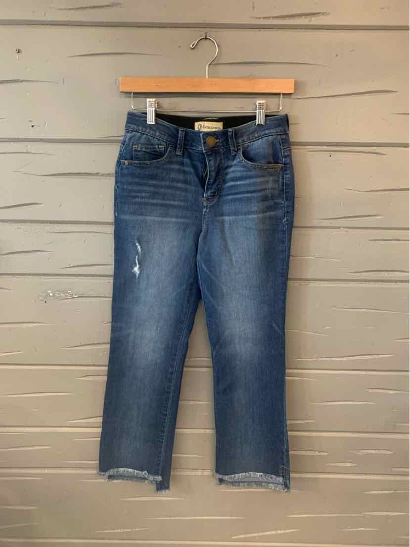 Blue cropped jeans with distressed details and frayed hems hanging on wooden hanger against beige wooden wall.