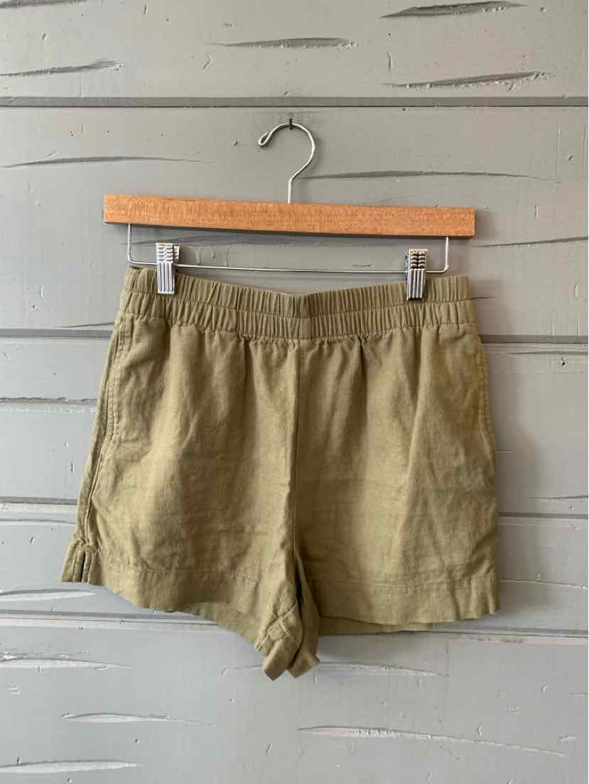 Light brown casual shorts with elastic waistband and side pockets hanging on wooden hanger against gray wooden wall.