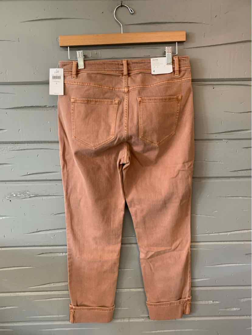 Light brown cropped denim jeans with rolled cuffs and back pockets hanging on wooden hanger against gray wooden wall.