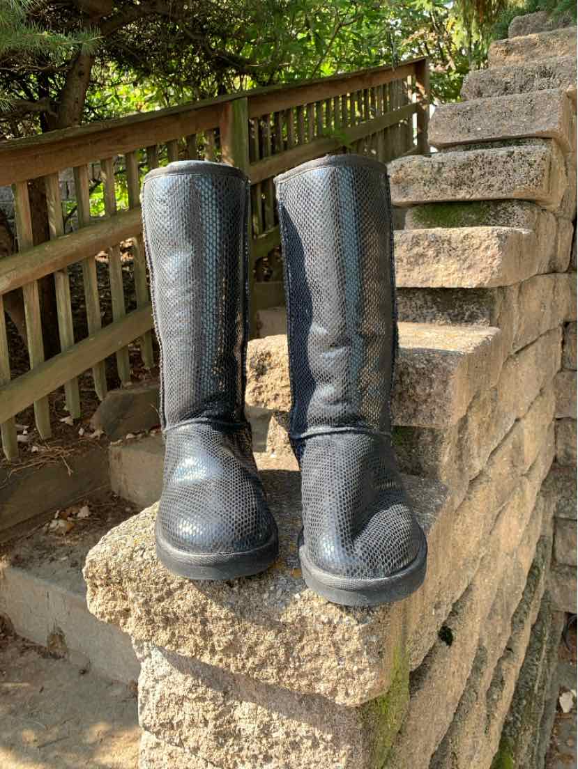 Pair of tall black UGG boots with rounded toes, suede-like finish, and subtle snake-print texture, displayed on a stone.