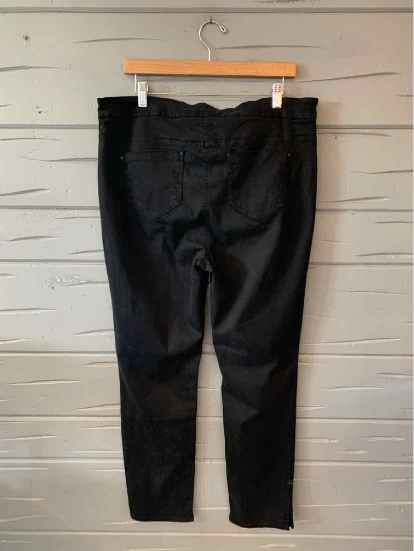 Black high-waisted slim-fit pants with belt loops and back pockets hanging on wooden hanger against a white wooden wall.