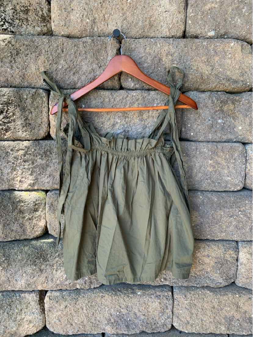 Olive green sleeveless top with gathered neckline and adjustable tie straps hanging on wooden hanger against stone wall.