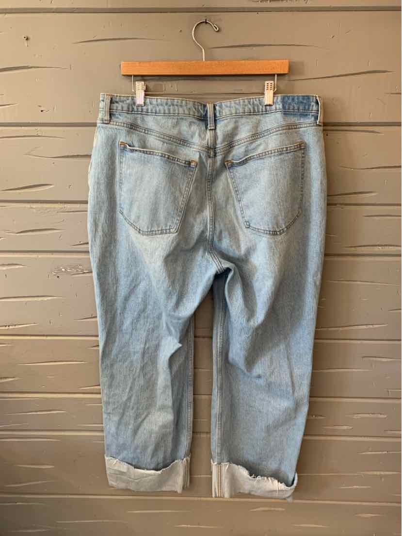 Light blue denim jeans with rolled cuffs and back pockets, hanging on a wooden hanger against a beige wooden wall.