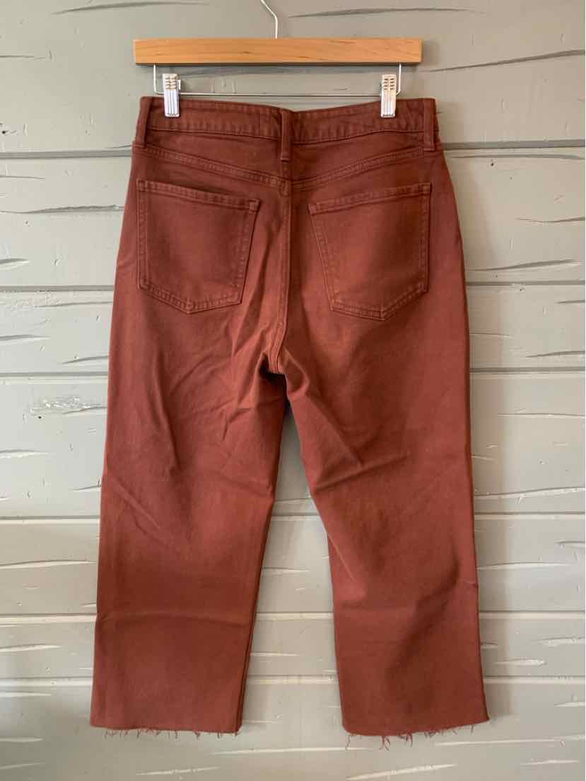 Rust-colored cropped pants with frayed hems and two back pockets hanging on wooden hanger.