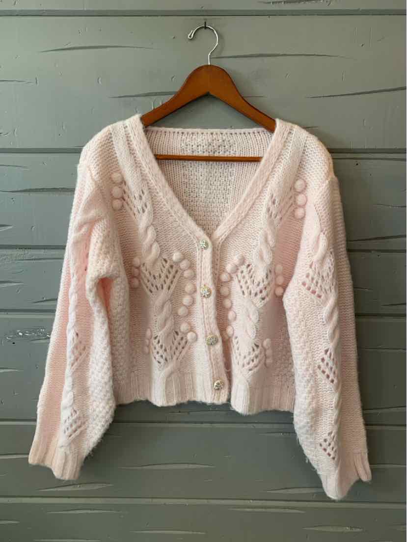 Pale pink chunky-knit cardigan with a V-neck, button front, and openwork floral patterns on a wooden hanger.