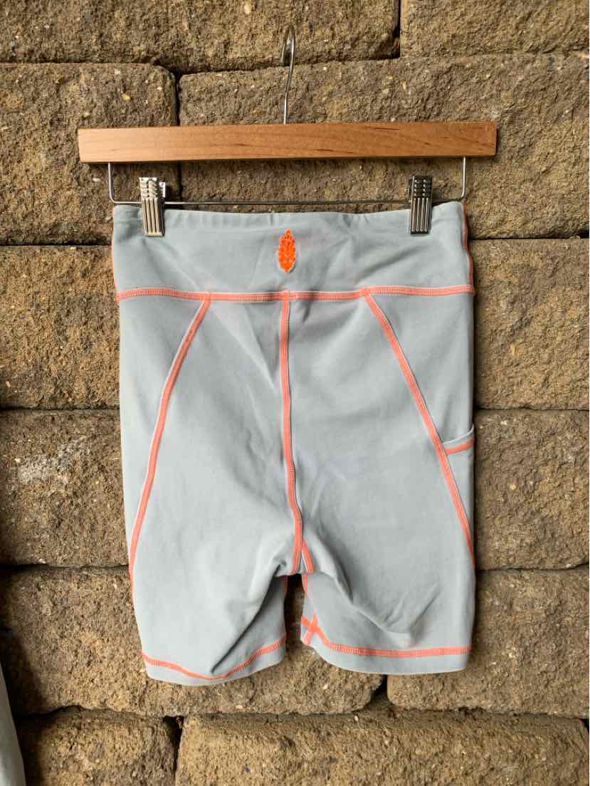 Light blue athletic shorts with orange seam accents and a high waistband hanging on a wooden hanger against a stone wall.