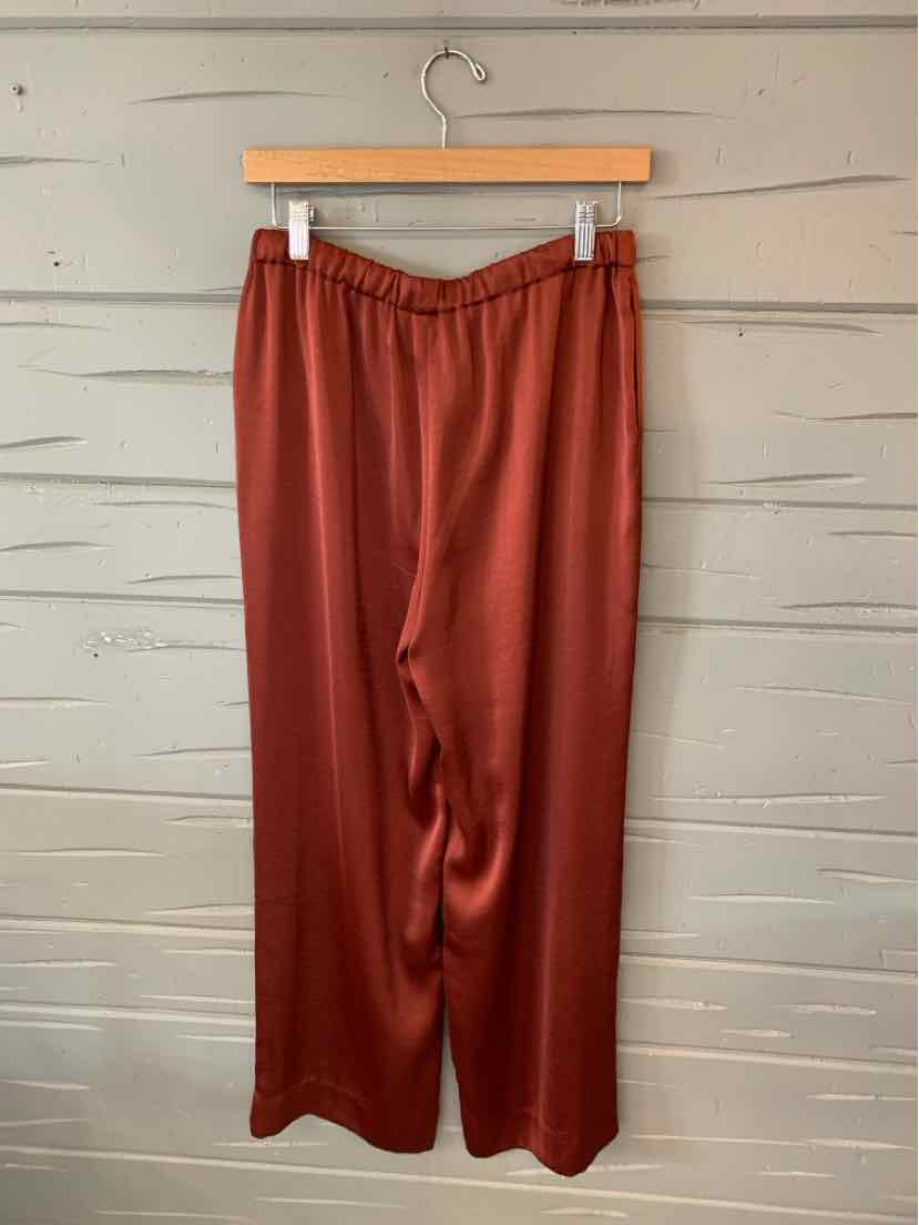 Brick-red wide-leg pants with elastic waistband hanging on wooden hanger against gray wooden wall.