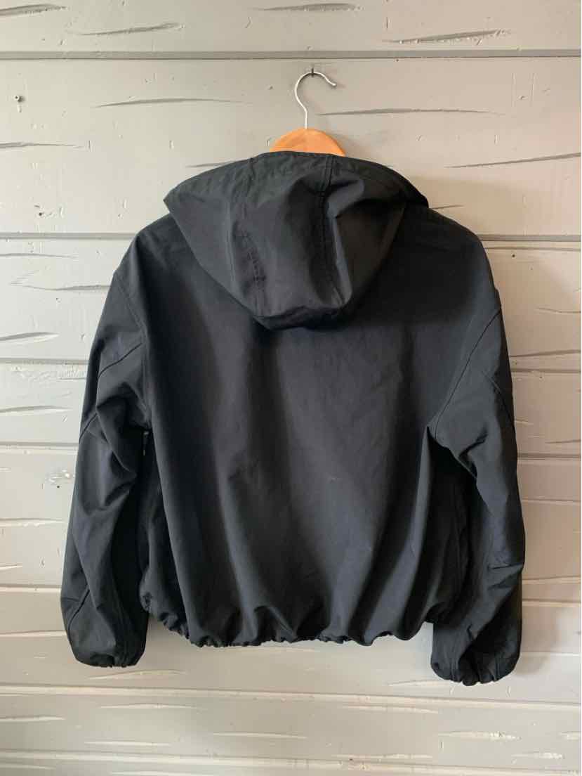 Black lightweight hooded jacket with elastic cuffs and hem hanging on wooden hanger against white wooden wall.